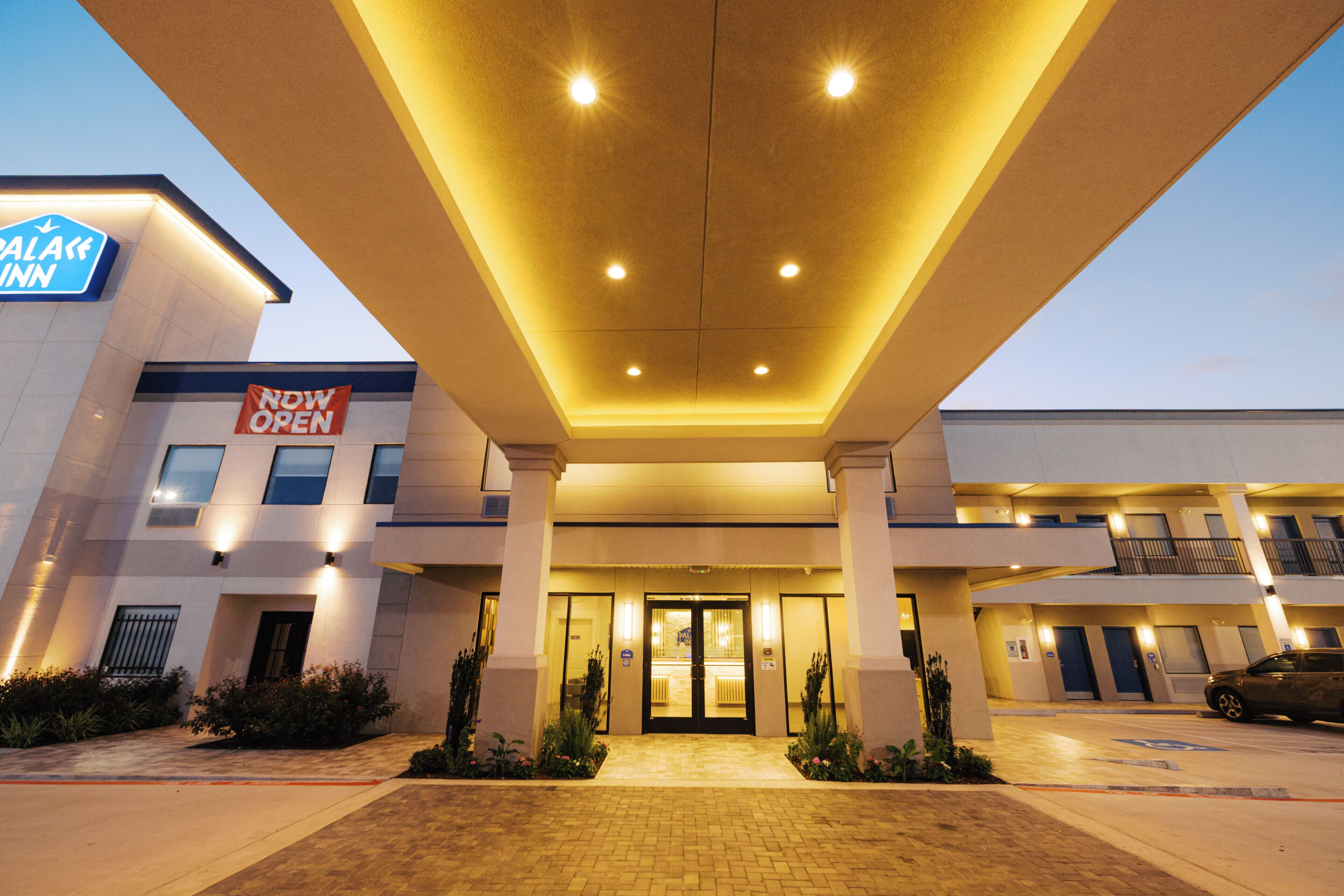 A hotel entrance with a 'NOW OPEN' sign, a parking lot, and a building with a 'PALACE INN' sign.
