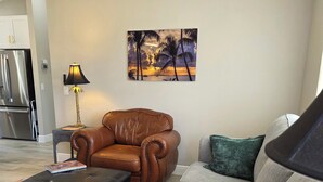 A living room with a brown leather chair, a grey sofa, and a black ottoman. There is a painting of a beach scene on the wall. - Cozy 2-bedroom house in The Villages Close To Golfing (Lady Lake)