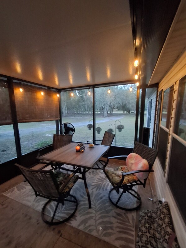 Un patio cubierto con mesa y sillas, ventanas amplias y luces colgantes.