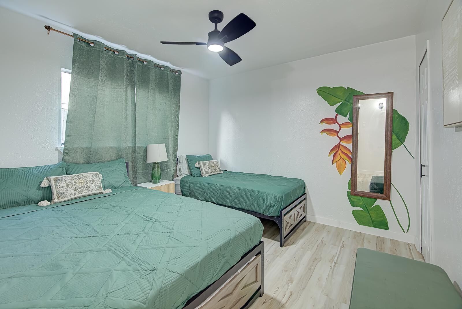 Una camera da letto con due letti, una ventola a soffitto, una finestra con tende e una porta con specchio e decorazione a foglia.