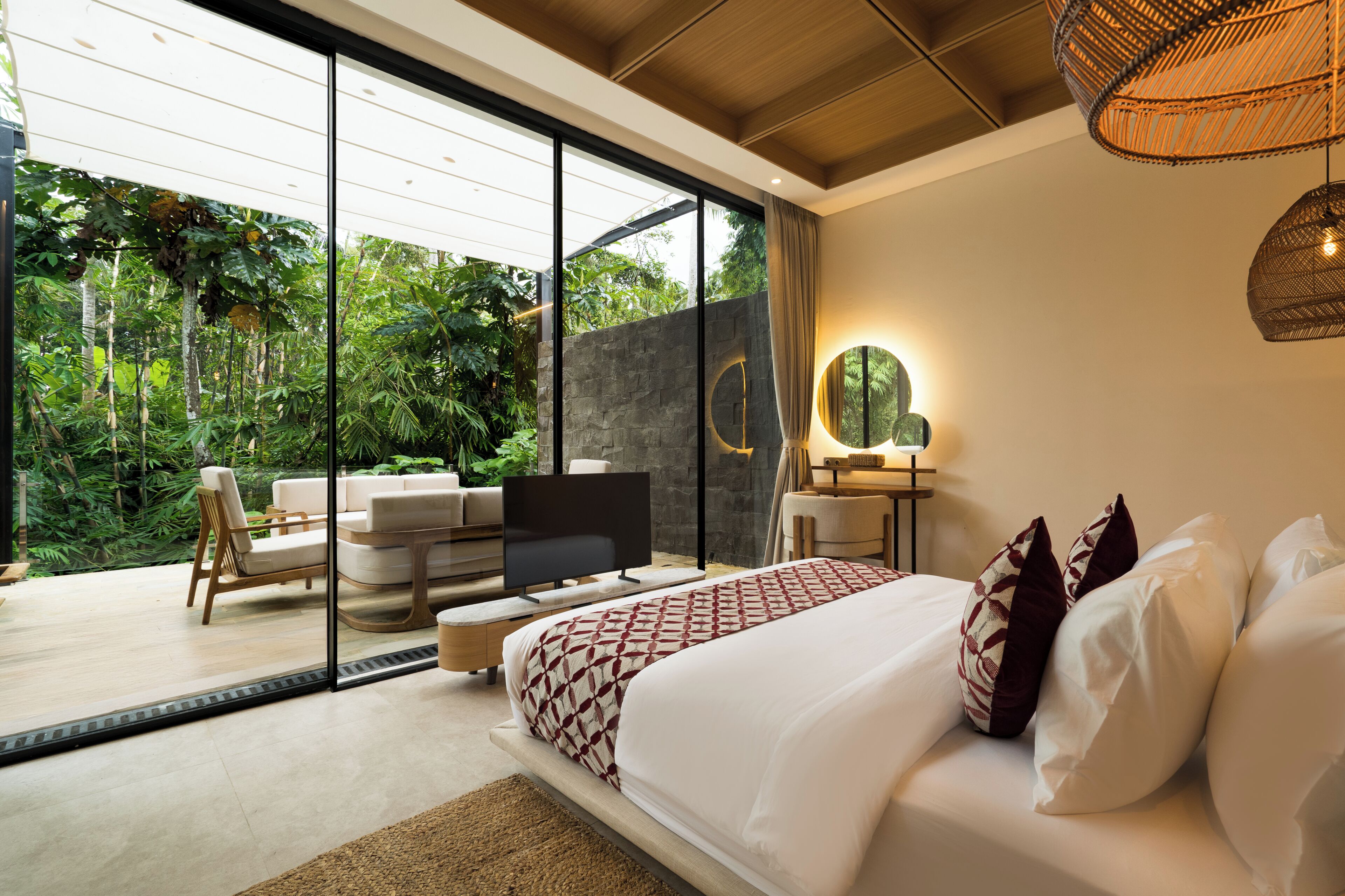 A bedroom with a large bed, a television, a sofa, and a dining area with a view of lush greenery.