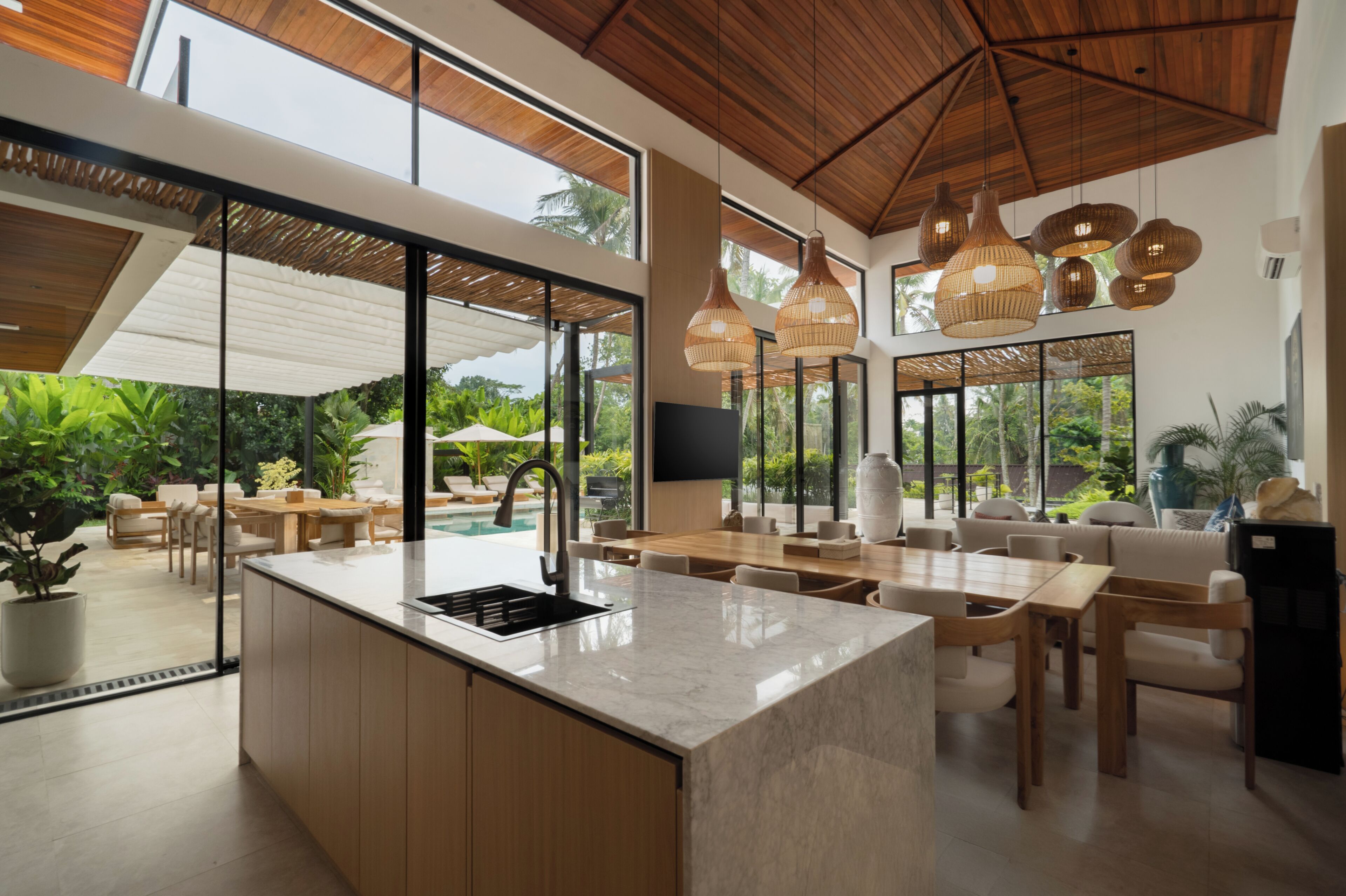 Een moderne keuken met een groot eiland, een eetgedeelte met houten meubels en grote ramen met uitzicht op groen.