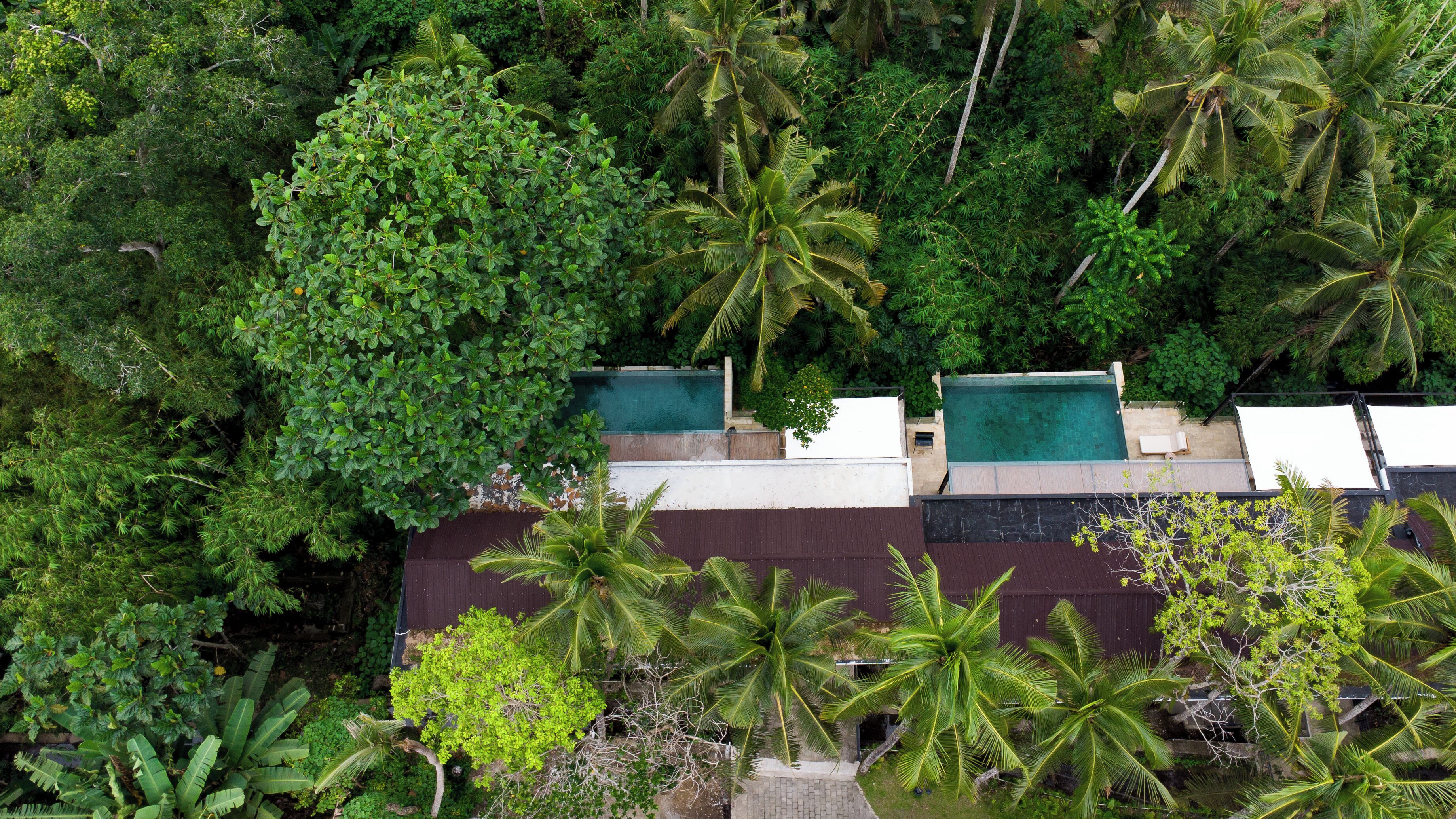 An aerial view of a resort with dense tropical vegetation, two swimming pools, and a large building with a dark roof.