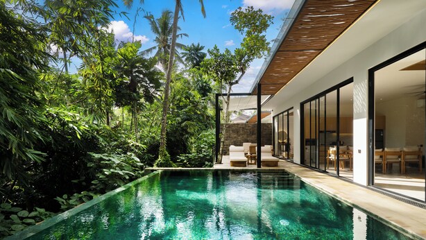 Una piscina moderna con agua cristalina, rodeada de exuberante vegetación y una casa contemporánea con amplias puertas de vidrio.