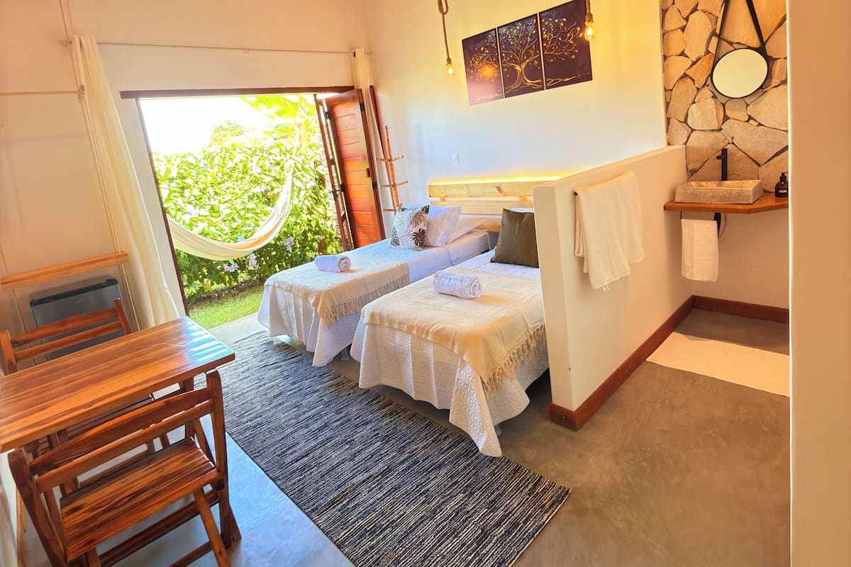 A room with a wooden ceiling, two beds with white linens, a small table, and a bench.