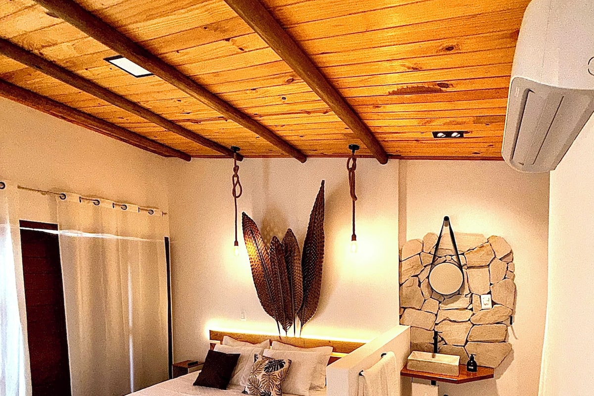 A bedroom with a wooden ceiling, a bed with a headboard, a nightstand, a stone wall, and a hanging light fixture.