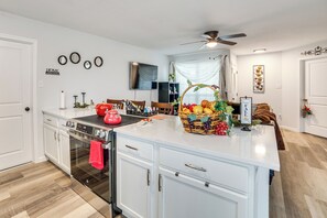 Una cocina moderna con armarios blancos, horno empotrado, hervidor rojo y una cesta de frutas sobre la encimera.