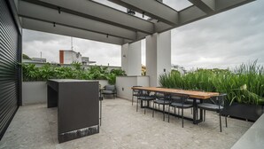 A rooftop terrace with a long wooden table and chairs, surrounded by potted plants and a modern architectural design.
