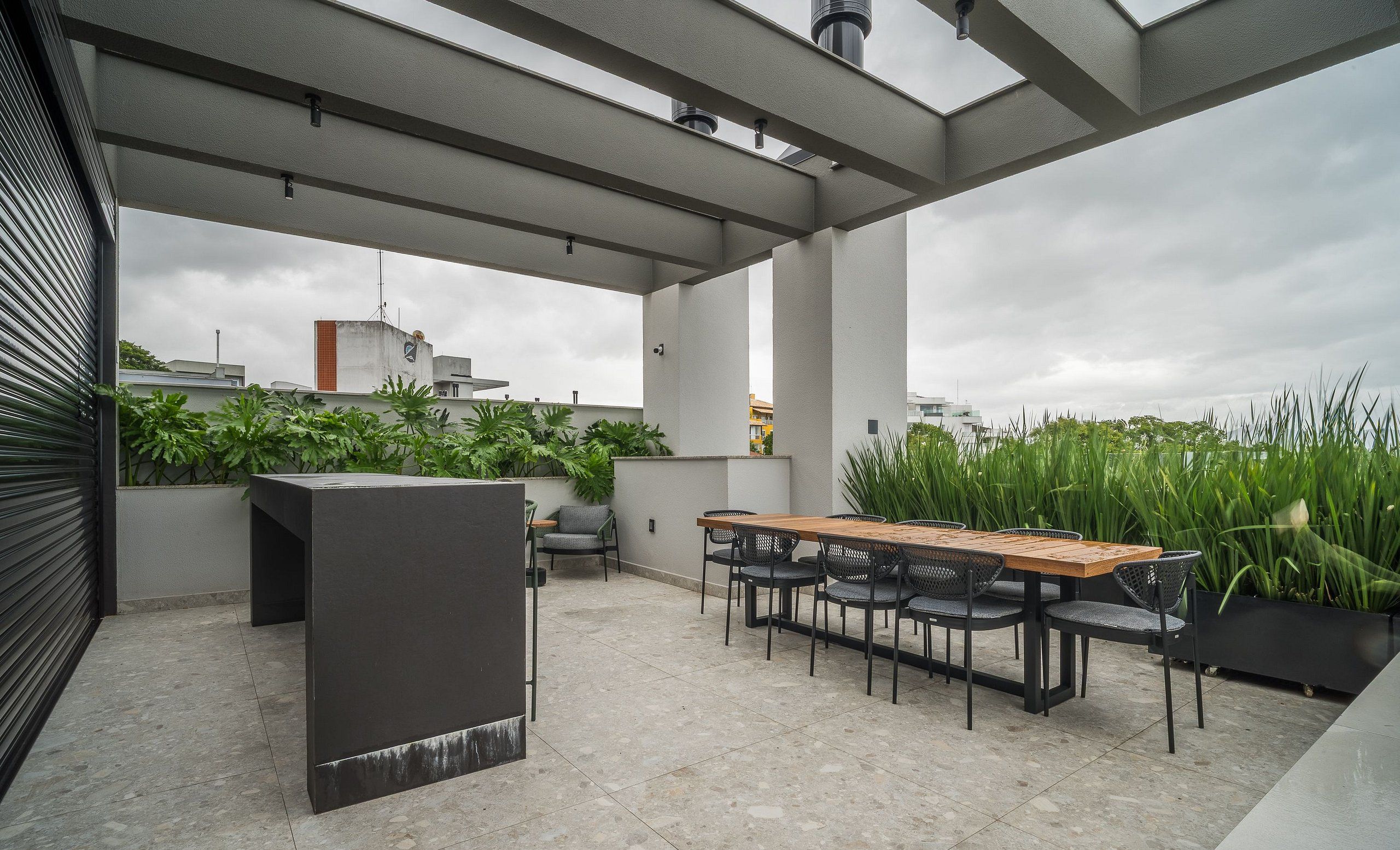Una terraza en la azotea con una larga mesa de madera y sillas, rodeada de plantas en macetas y un diseño arquitectónico moderno.