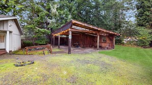 A rustic wooden cabin with a porch and string lights, surrounded by a well-maintained lawn and dense trees. - Charming house w/ wifi, fireplace & dog-friendly (Shelton)
