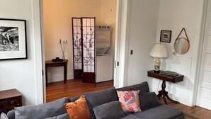 Un salon avec un canapé gris, un pouf et une petite table basse. On y voit une lampe, un miroir et un tableau encadré au mur. Un meuble en bois et une petite table avec un vase sont également visibles.