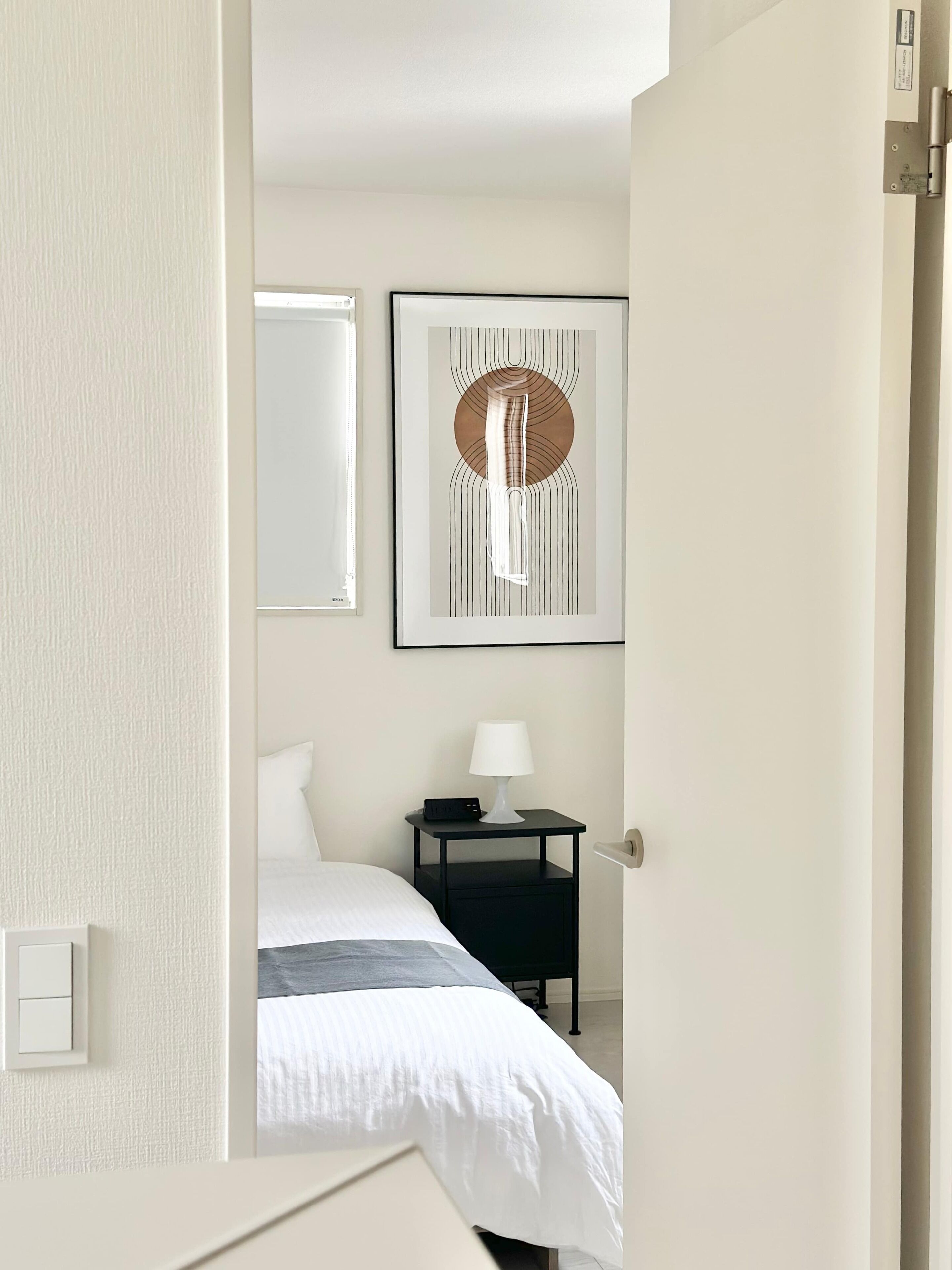 A hotel room with a bed, bedside table with a lamp, a framed abstract artwork, and a window.