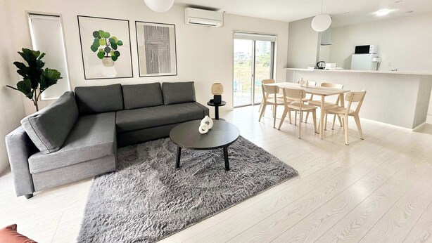 A modern living room with a grey sofa, a round coffee table, and a dining area with a white table and chairs. - 10 mins to Kumamoto Airport Premium 4BR Retreat by Restavio (Ozu)