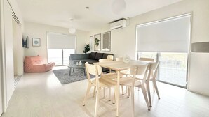 A modern living room with a dining area, a grey sofa, a pink bean bag, and a large window with blinds. - 10 mins to Kumamoto Airport Premium 4BR Retreat by Restavio (Ozu)
