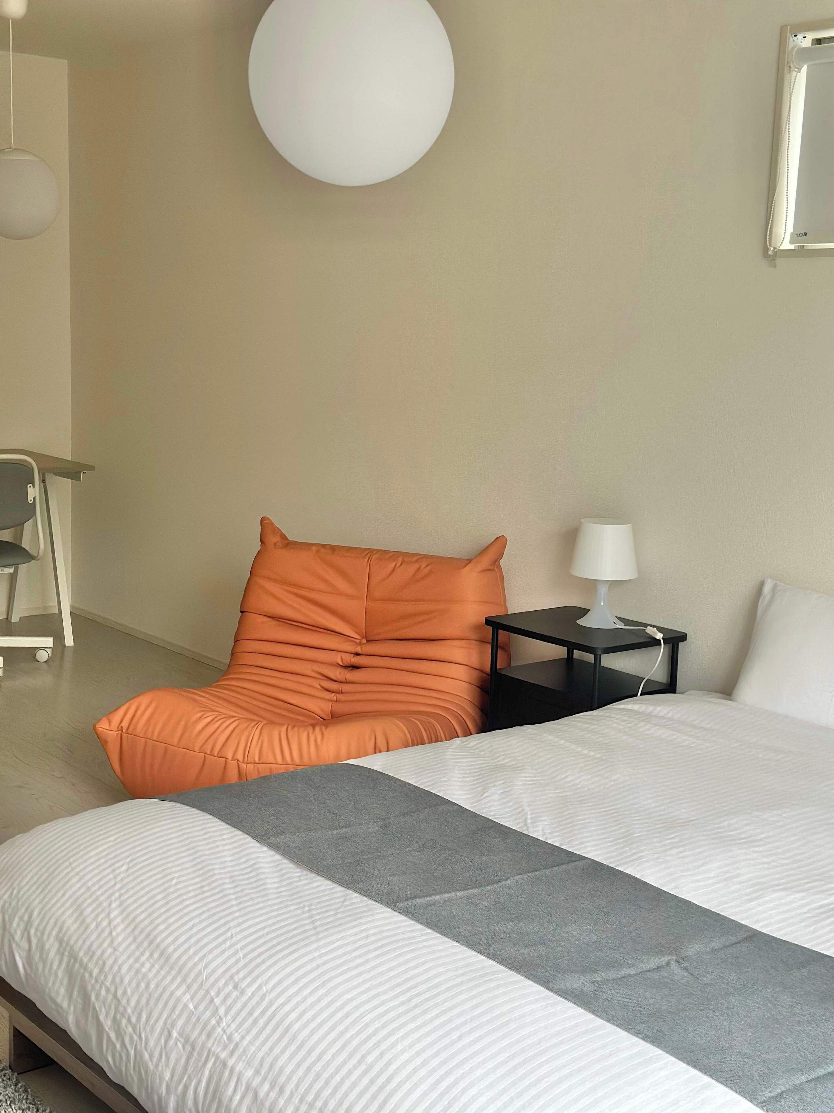 Una habitación de hotel moderna con una cama, un sillón naranja, una mesita de noche con una lámpara y un gran aplique redondo en la pared.