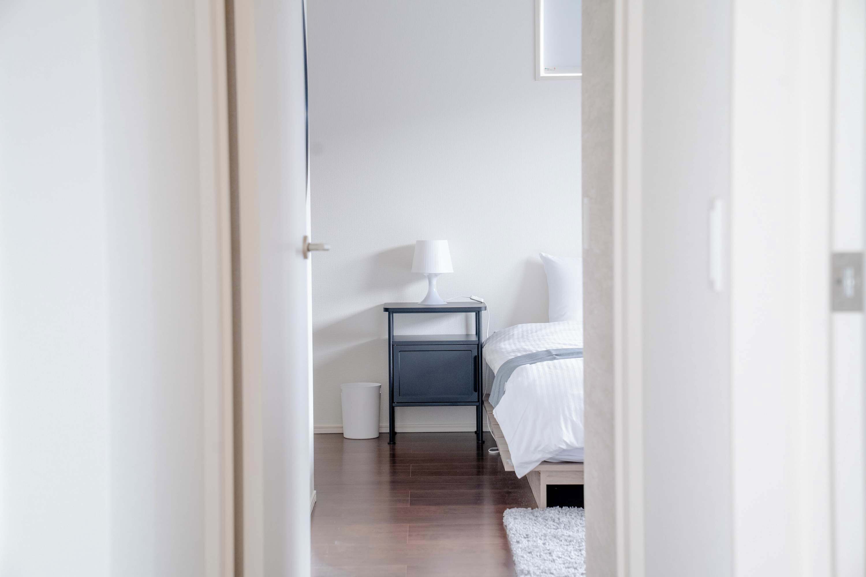 Une chambre à coucher avec un lit, une table de chevet avec une lampe, un tapis blanc et une poubelle blanche.