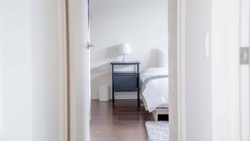 Une chambre à coucher avec un lit, une table de chevet avec une lampe, un tapis blanc et une poubelle blanche.