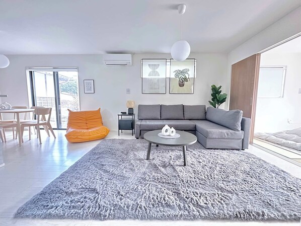 A modern living room with a grey sofa, a round coffee table, and a large grey rug. There is a small orange bean bag chair and a dining table with chairs. The room has large windows and a plant on the windowsill. - Near Airport and Aso Volcano Modern 4BR Residence by Restavio (Ozu)
