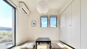 Et minimalistisk rum med tatamigulv, et bord af tatami-måtter, to tatami-måtter som siddepladser, en loftlampe og et stort vindue med udsigt.