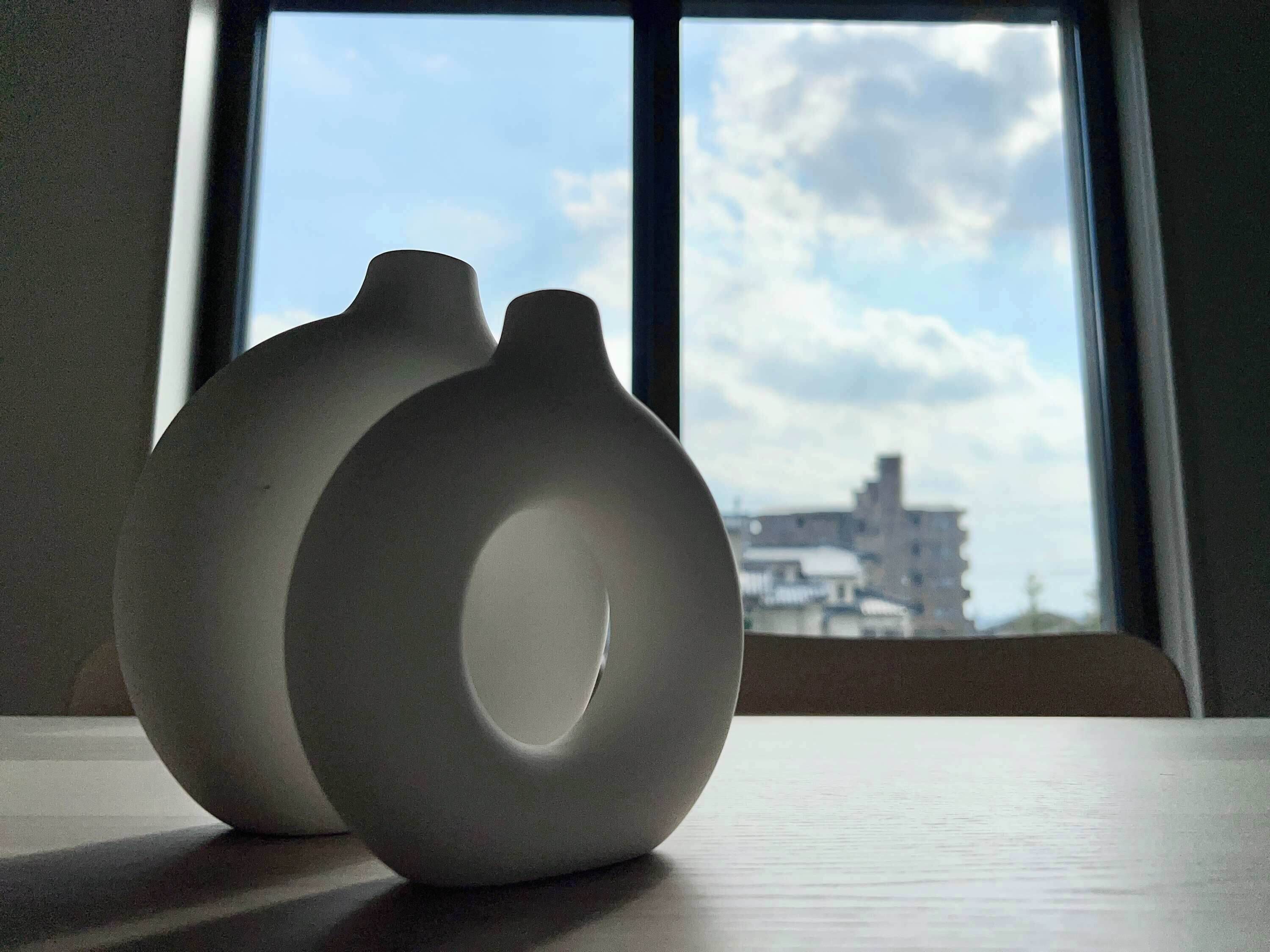 Deux vases en céramique blanche sont posés sur une table près d’une fenêtre, d’où l’on aperçoit des immeubles et un ciel nuageux.