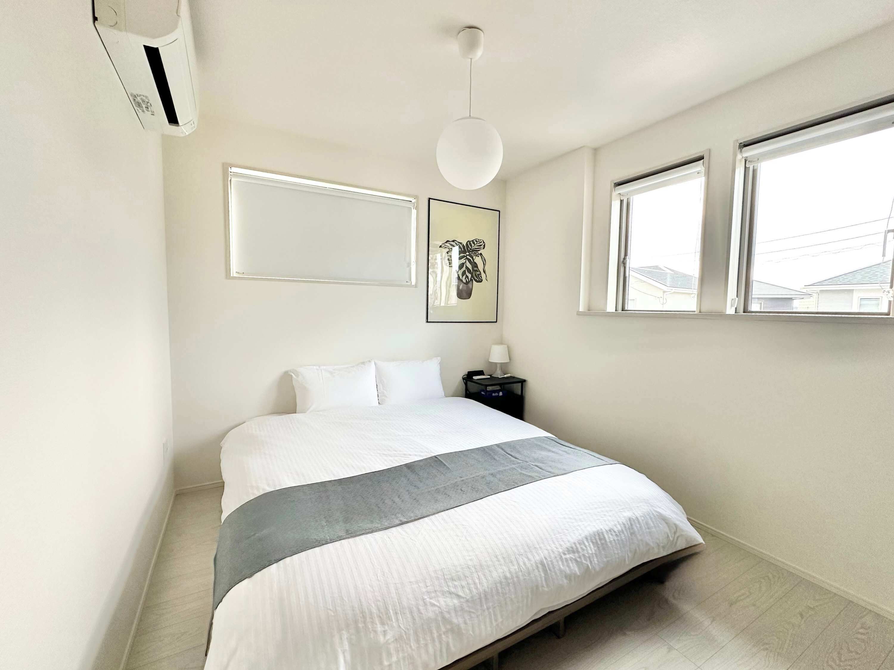 A bedroom with a bed, a bedside table with a lamp, a wall-mounted air conditioner, and a window with a view of houses.