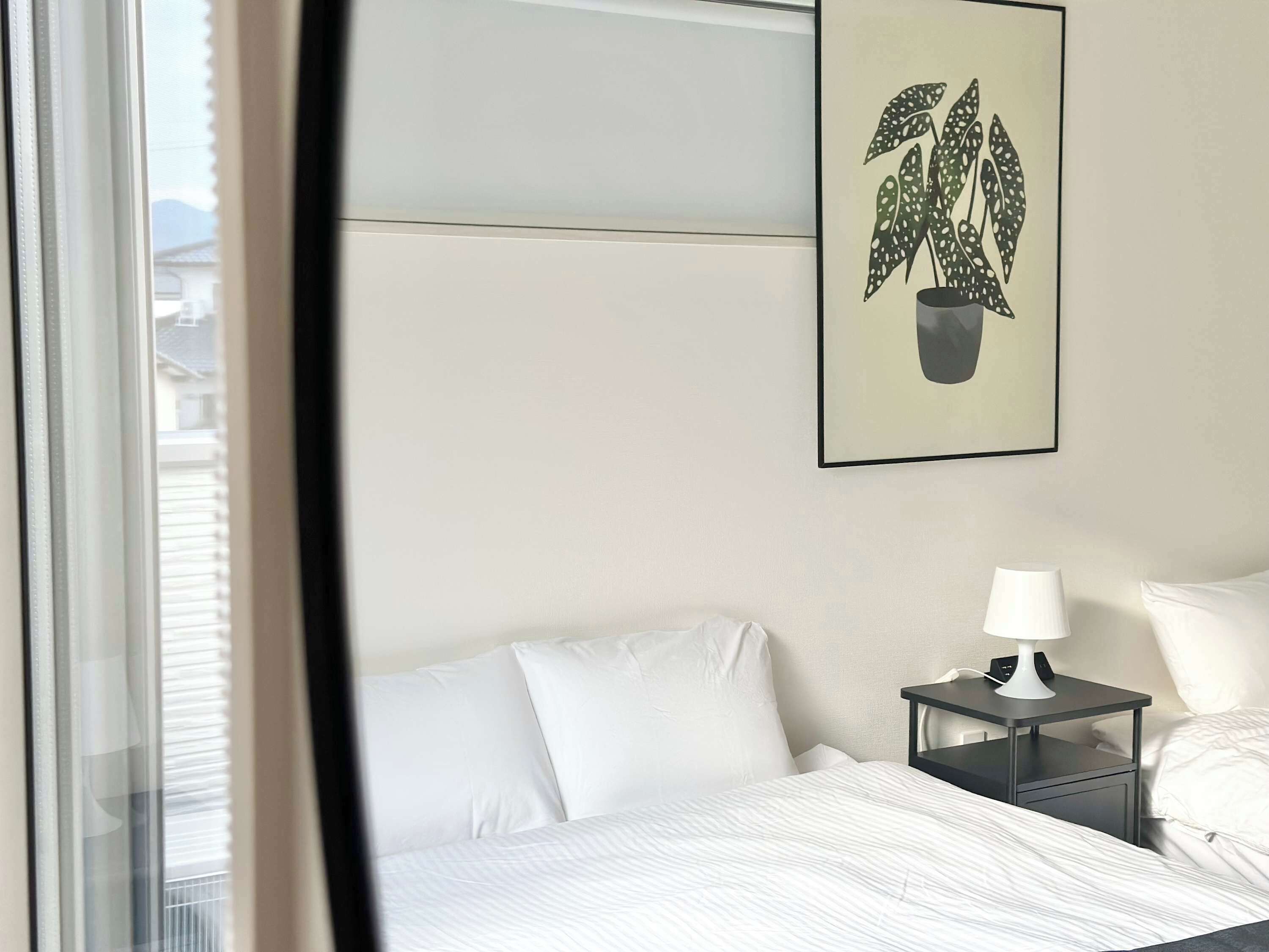A hotel room with a bed, white bedding, a nightstand with a lamp, and a framed plant on the wall.