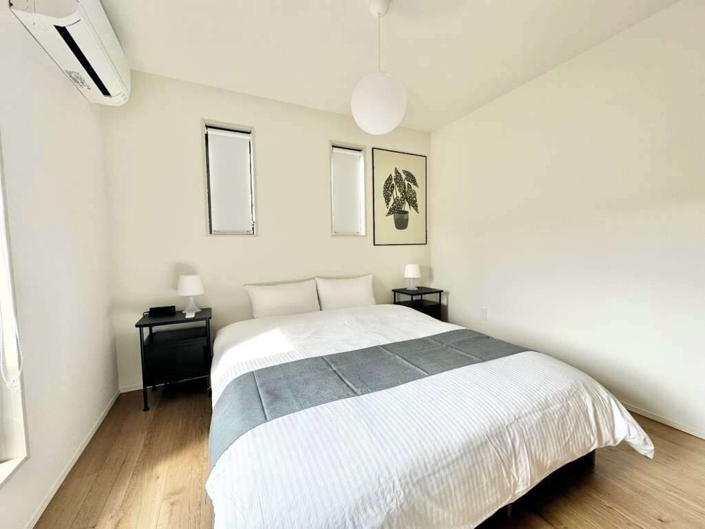 A bedroom with a bed, two bedside tables, a wall-mounted air conditioner, a window, and two framed pictures.