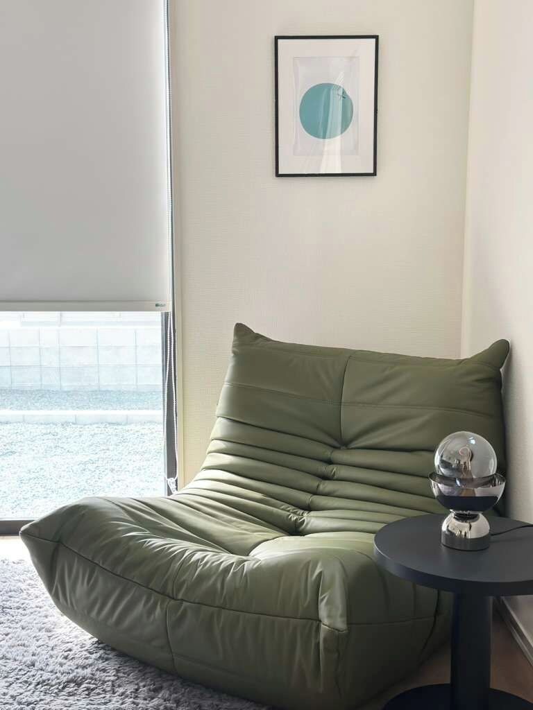 Uma poltrona verde com almofadas próxima a uma janela com vista para a praia, um quadro na parede e uma pequena mesa com um globo de cristal.