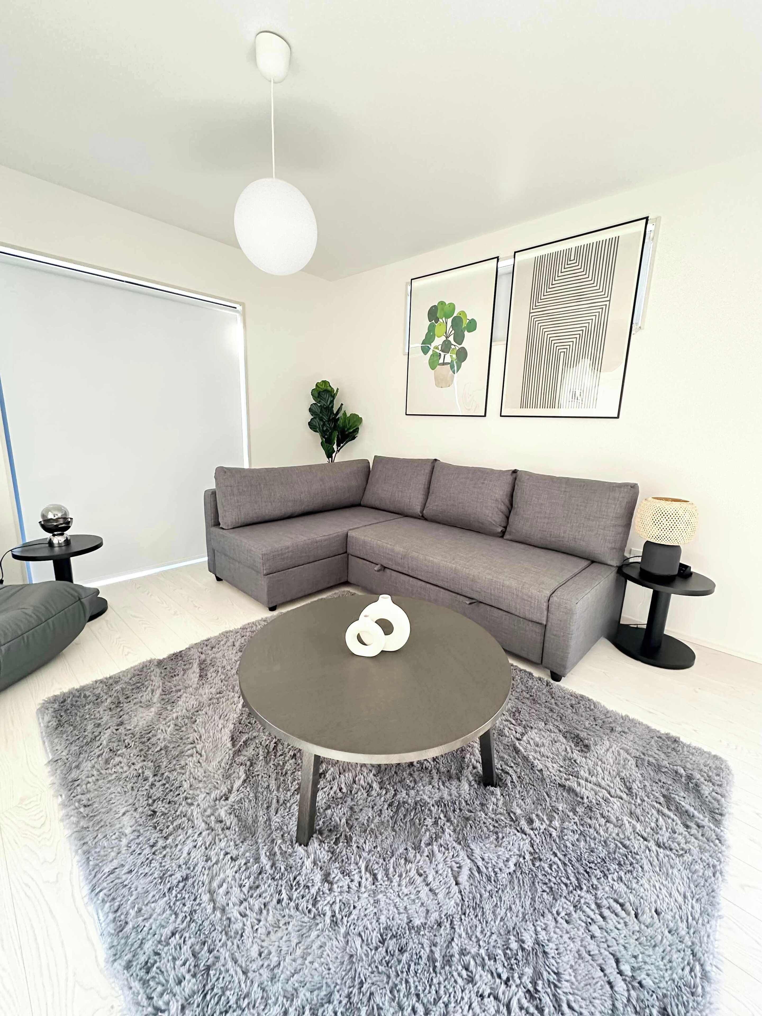 Een moderne woonkamer met een grijze hoekbank, een ronde salontafel en een zachtgrijs vloerkleed. Er hangen twee ingelijste kunstwerken aan de muur, staat een potplant en is er een klein bijzettafeltje met een lamp.