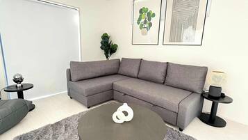 Een moderne woonkamer met een grijze hoekbank, een ronde salontafel en een zachtgrijs vloerkleed. Er hangen twee ingelijste kunstwerken aan de muur, staat een potplant en is er een klein bijzettafeltje met een lamp.