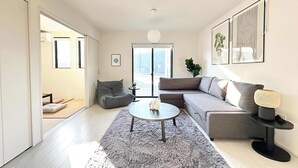 A modern living room with a grey sofa, a round coffee table, and a grey shag rug. There is a large window with a view of buildings outside. - 10 mins to kumamoto airport Spacious 4BR House by Restavio (Ozu)