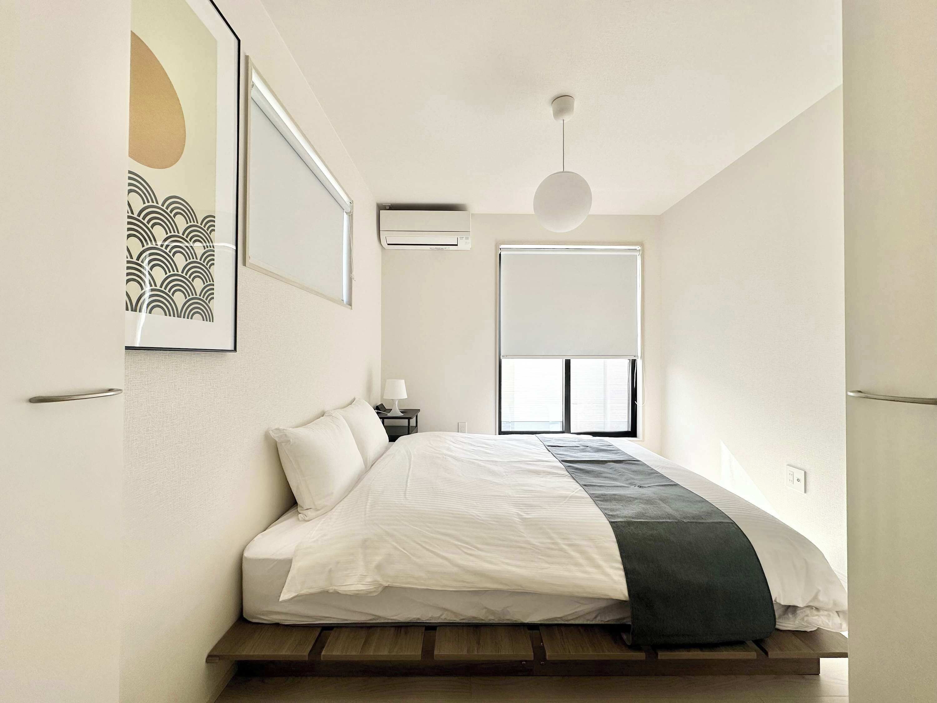 Et minimalistisk soveværelse med seng, natbord, lampe, et vindue med skodder og en loftslampe.