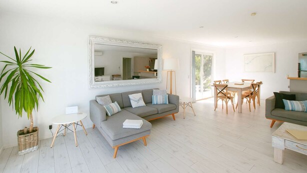 Una sala de estar moderna con un sofá gris, una mesa de comedor con sillas, un espejo grande y una planta en maceta.