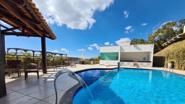 A swimming pool with a pergola and lounge chairs, a modern building with a green-tiled facade, and a clear blue sky. - Complete two-story house in Escarpas do Lago – Comfort and Leisure: Your Dream Retreat (Capitólio)