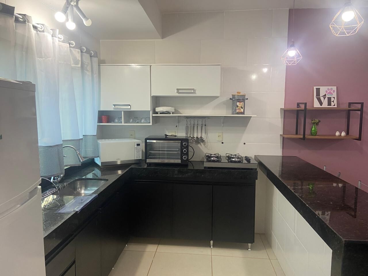 Una cocina moderna con encimeras negras, armarios blancos y un horno empotrado. Hay un lavamanos con un grifo, un tostador y una pequeña estantería con objetos decorativos.