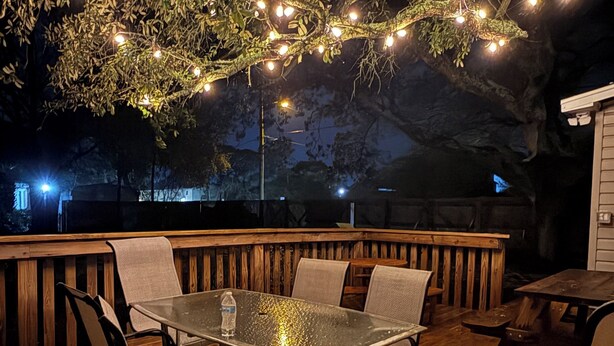 Eine Terrasse im Freien mit einem Tisch und Stühlen mit Glasplatte, umgeben von Holzgeländern und Lichterketten.