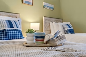 A neatly made bed with striped pillows, a small potted plant, a striped mug, and an open book on a tray. - River Oaks Condo near beach & golf. Pool, tennis & free parking (Myrtle Beach)