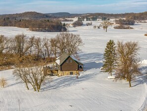 Eine Holzhütte mit Steinunterbau und einem grünen Dach, umgeben von schneebedeckten Feldern und vereinzelten Bäumen.