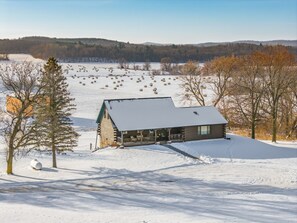 Une cabane en rondins dont le toit est recouvert de neige, entourée d’un paysage enneigé parsemé de bottes de foin et d’arbres.