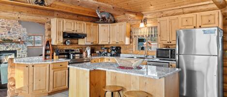 A spacious kitchen with wooden cabinets, a large island with stools, a stainless steel refrigerator, and a stone fireplace.