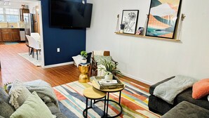 Un salon avec un canapé, une table basse et un téléviseur fixé au mur. On aperçoit une cuisine au fond de la pièce.