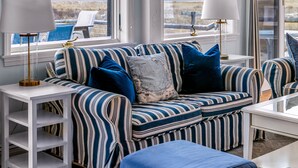 Ein modernes Wohnzimmer mit einem gestreiften Sofa, zwei Beistelltischen mit Lampen, einem weißen Couchtisch und großen Fenstern, die einen Blick ins Freie bieten.