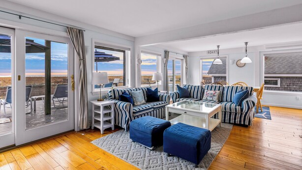 Ein geräumiges Wohnzimmer mit einer großen Schiebeglaswand, die auf eine Terrassenausläufe führt, einer gestreiften Sofagarnitur, einem Couchtisch und Blick auf die Landschaft.