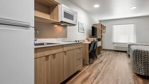 A compact kitchen with wooden cabinets, a microwave, and a sink. There is a small dining area with a chair and a table. - Woodspring Suites Lancaster North (Lancaster)