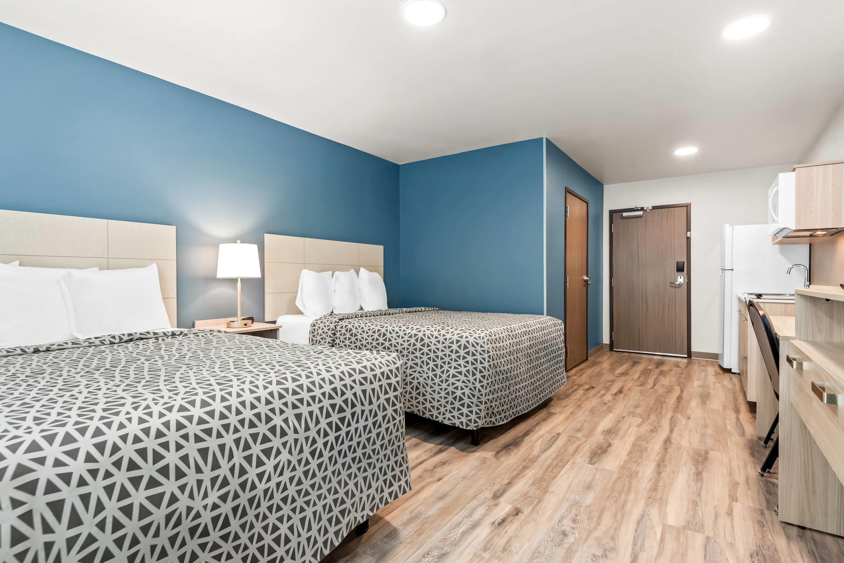 A hotel room with two beds, a desk, a lamp, a door, and a kitchenette.