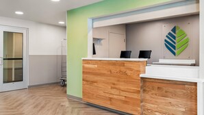 A reception desk with a wooden finish, a green wall, and a logo on the wall. - Woodspring Suites Lancaster North (Lancaster)
