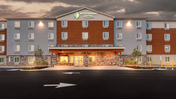 A multi-story hotel with a stone facade, a sign reading 'Woodspring Suites,' and a parking lot with directional arrows. - Woodspring Suites Lancaster North (Lancaster)