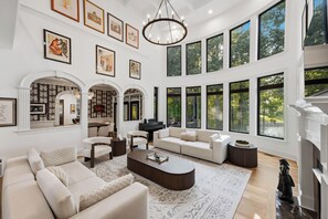 A spacious living room with a piano, comfortable seating, and large windows offering views of the outdoors. - Spacious Lake Norman Home • Hot Tub + Firepit + Deck | Sleeps 22 | Lion Manor (Denver)