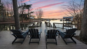 A wooden deck with four Adirondack chairs facing a calm lake at sunset. - Lake Norman Estate • Hot Tub + Fire Pit + Movie Theater | Lion Manor (Denver)