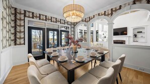 A dining room with a large table set for six, surrounded by upholstered chairs. A chandelier hangs from the ceiling, and the room features white walls and wooden flooring. - Lake Norman Estate • Hot Tub + Fire Pit + Movie Theater | Lion Manor (Denver)